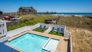 A pool area with lounge chairs, a wooden deck, and a view of the beach and pier.