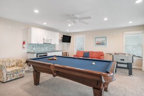 Een pooltafel met een keu, een bank, een tafelvoetbalspel en een keukenhoek met kastjes en een magnetron.