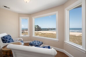 A room with a view of a beach and pier, featuring a large window, a chair, and a small table.
