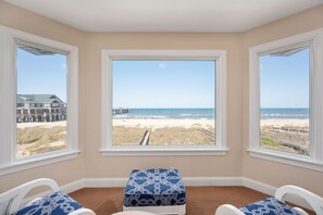 A room with three large windows offering a beach view, a carpeted floor, and two white chairs with blue cushions.