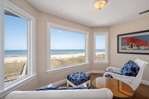 A room with large windows overlooking a beach, a white chair with a patterned cushion, and a framed artwork on the wall.