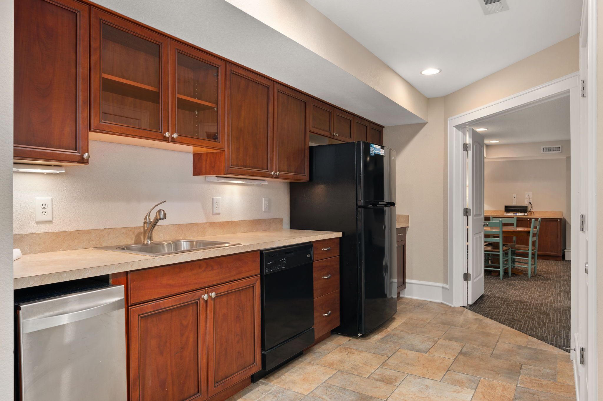 A modern kitchen with wooden cabinets, a stainless steel dishwasher, a black refrigerator, and a tiled floor.