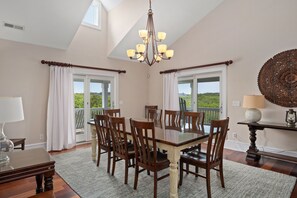 A dining room with a large table, chairs, a chandelier, and a view through the windows.