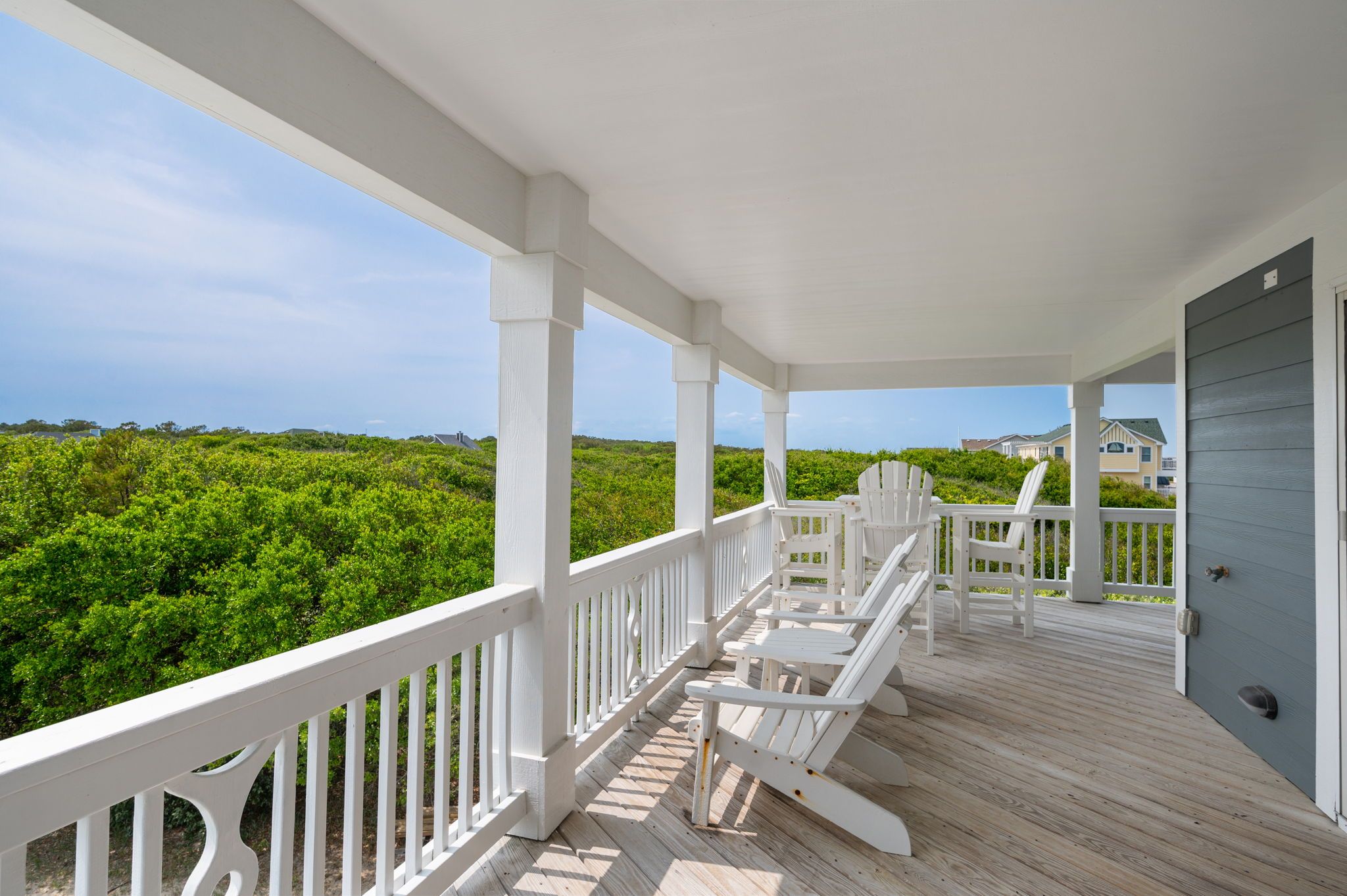 A covered porch with white railings and Adirondack chairs, overlooking a lush green landscape.