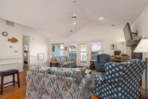 A spacious living area with patterned sofas, a wooden floor, a ceiling fan, and a television.