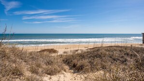 A beach with a clear sky, sandy dunes, and a wooden structure in the distance. - Nags Head 225:Historic oceanfront, sandy path to the beach, comfy & cozy (Nags Head)