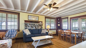 A room with a wooden floor, a blue sofa, a dining table with chairs, a ceiling fan, and a stone fireplace. - Nags Head 225:Historic oceanfront, sandy path to the beach, comfy & cozy (Nags Head)
