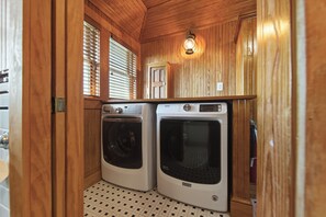 A compact laundry room with two front-loading washing machines, wooden walls, a small window with blinds, and a wall-mounted light fixture.