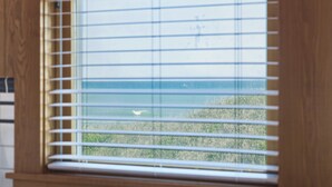 A window with horizontal blinds, a view of the sea, and some greenery outside. - Nags Head 225:Historic oceanfront, sandy path to the beach, comfy & cozy (Nags Head)