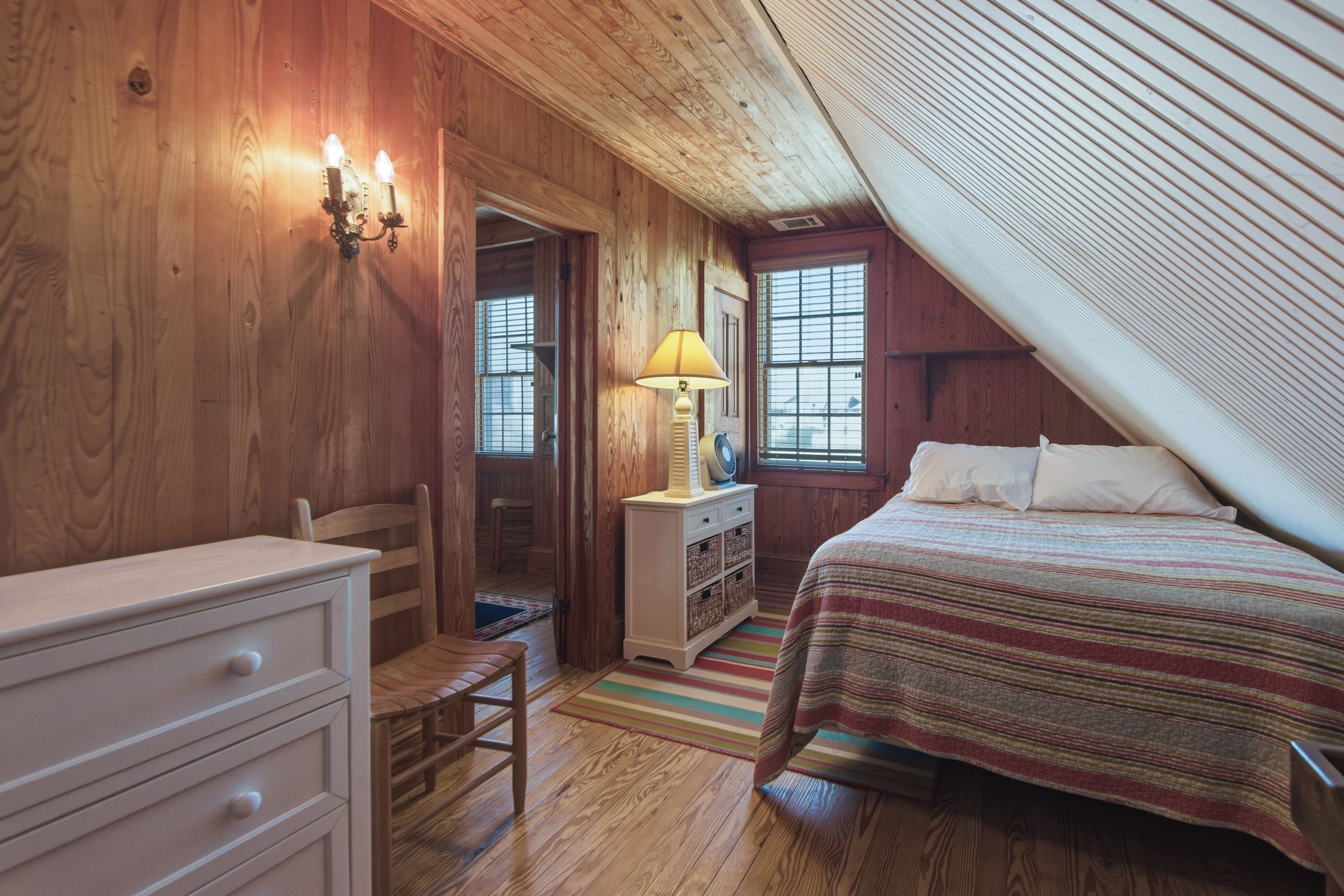 A bedroom with a wooden ceiling, a bed with a striped bedspread, a nightstand with a lamp, and a dresser.