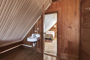 Un interior de madera con techo inclinado, un lavamanos blanco y un dormitorio visible con cama y alfombra estampada.