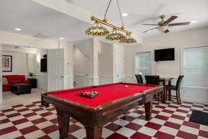 A pool table with a red felt surface, a ceiling fan, and a chandelier with stained glass lampshades.