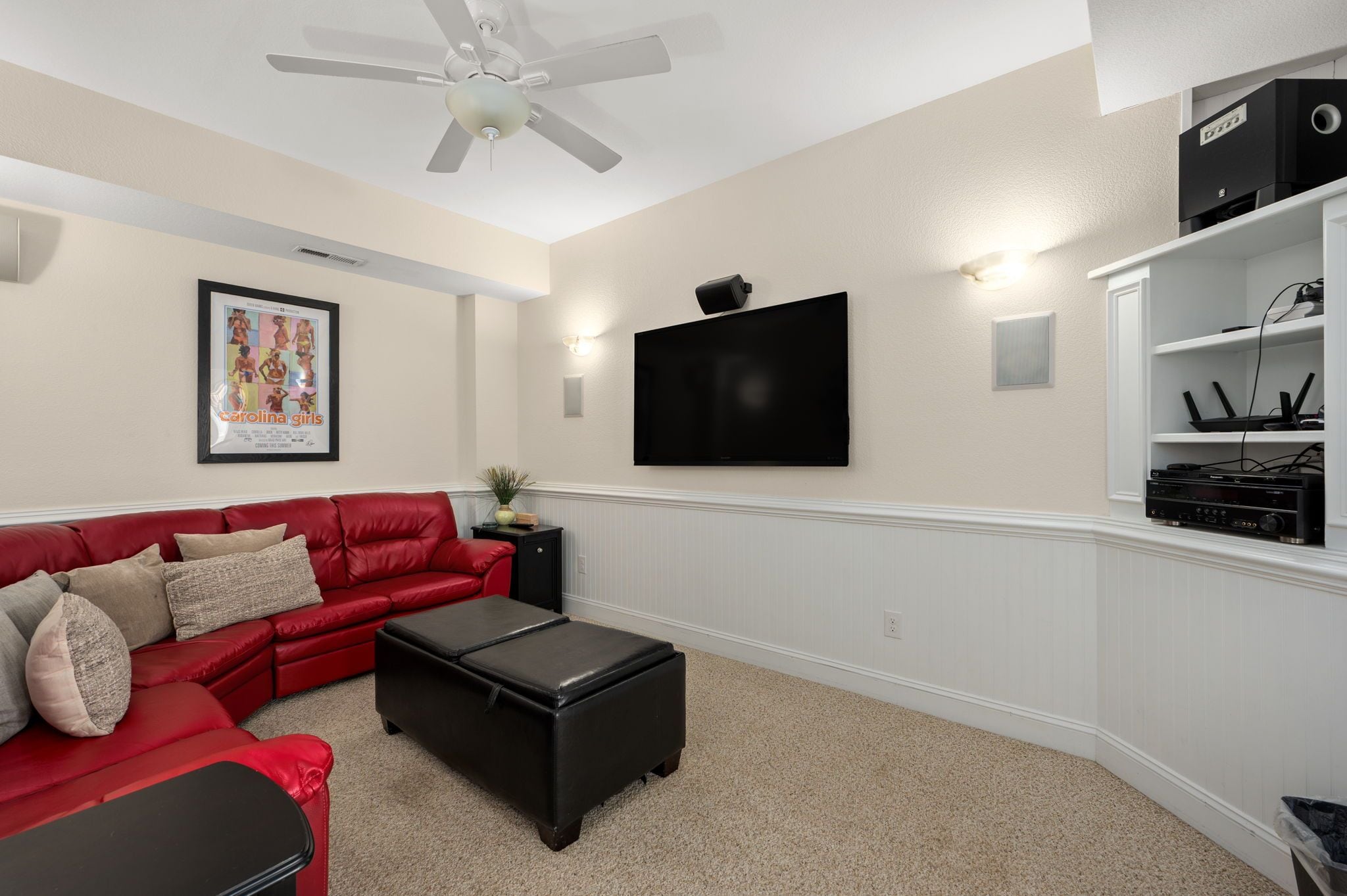 A living room with a red sofa, a black ottoman, a ceiling fan, a flat-screen TV, and a white entertainment unit with electronic devices.