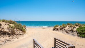 Une plage avec une promenade en bois menant à l’eau, des dunes de sable et un ciel bleu clair.