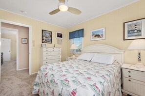 A bedroom with a bed, a dresser, a nightstand, a ceiling fan, and wall art. - L'il Seabreeze-Oceanfront, ocean views, awesome sunrises, steps to the beach! (Nags Head)