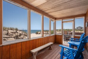 A covered outdoor seating area with wooden benches and chairs, large windows offering a view of the beach and dunes.