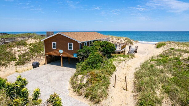 Une propriété située en bord de mer, comprenant une maison de plain-pied, une véranda et un chemin de sable menant à la plage.