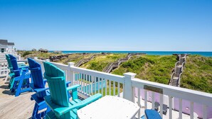 A deck with blue and white Adirondack chairs, a white table, and a view of the beach and ocean. - Celestial Shore-Oceanfront, pool, hot tub, elevator, rec room & dog friendly! (Nags Head)