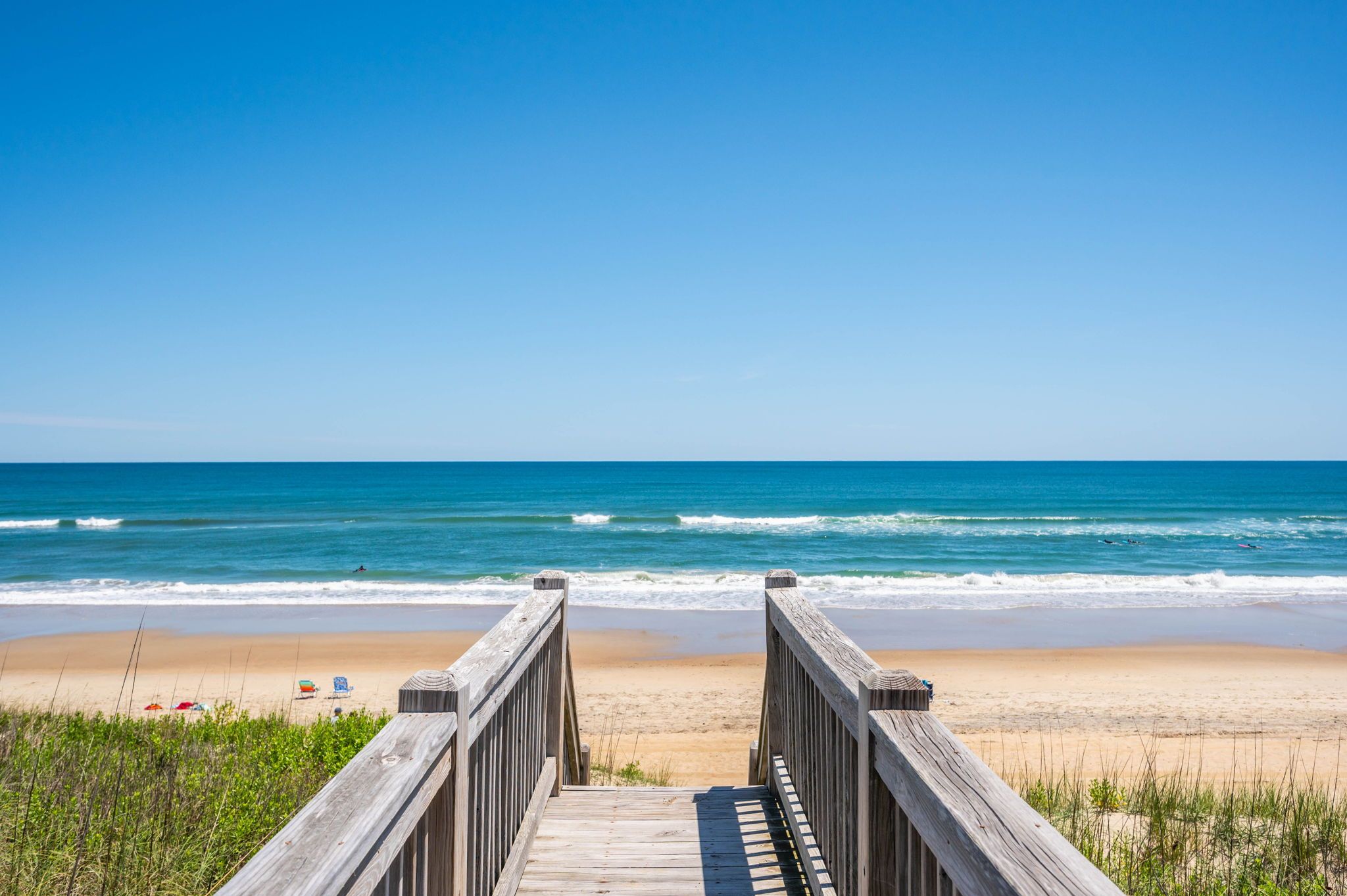 A wooden boardwalk leading to a beach with clear blue water and a sandy shore.