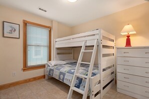 Un lit superposé avec une échelle, une fenêtre avec des stores, une commode avec des tiroirs et une lampe fixée au mur.