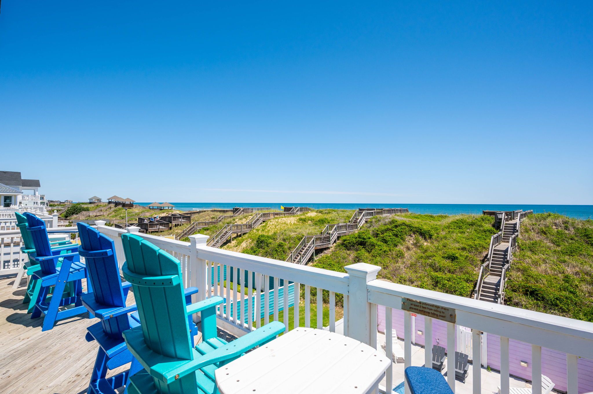 A deck with blue and white Adirondack chairs, a white table, and a view of the beach and ocean.
