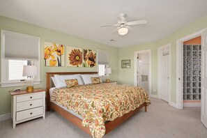 A bedroom with a bed, nightstand, lamp, ceiling fan, and floral artwork on the wall.