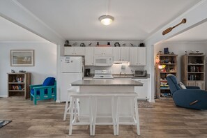 Una cocina moderna con armarios blancos, una isla central con taburetes, un microondas y una estufa. Hay una silla azul y una estantería con objetos decorativos.