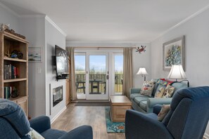 A living room with a fireplace, a television, a sofa, a coffee table, and a view of the outdoors through a glass door. - Pooh's Corner: Oceanfront condo & Patio, steps to the beach, dog friendly! (Nags Head)