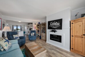 A living room with a sofa, armchair, and TV wall-mounted above a fireplace. - Pooh's Corner: Oceanfront condo & Patio, steps to the beach, dog friendly! (Nags Head)
