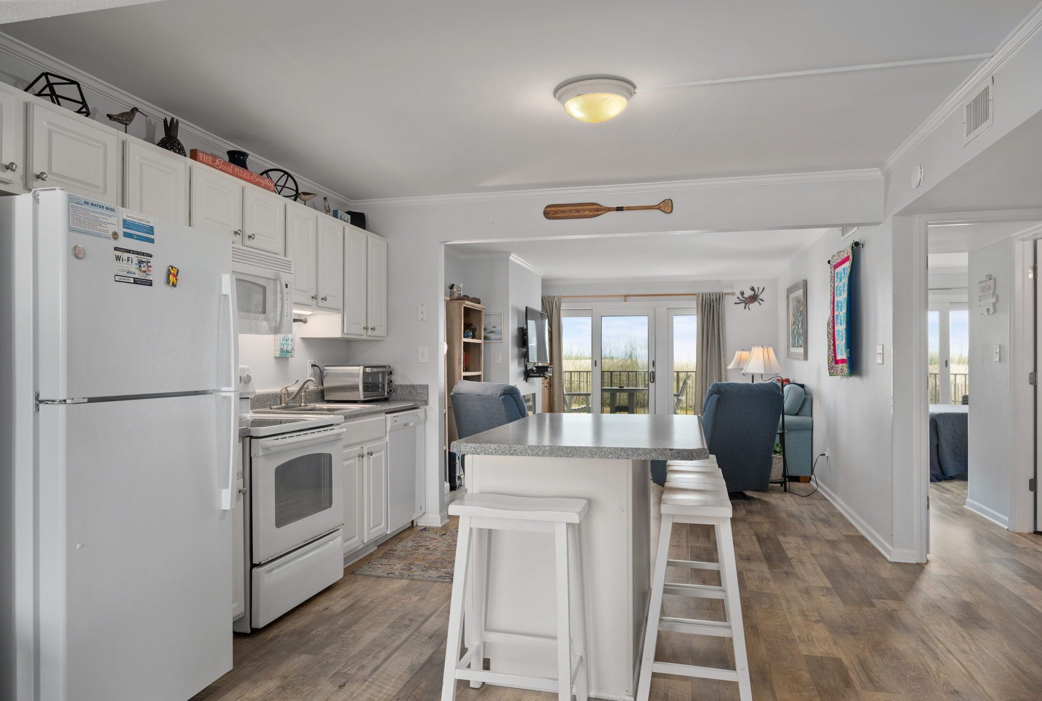 A modern kitchen with white cabinets, a built-in oven, and a microwave. There is a bar area with stools and a ceiling-mounted paddle.
