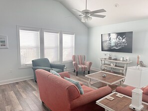 Un salon moderne avec un ventilateur de plafond, un grand téléviseur à écran plat, un fauteuil à rayures, un canapé bleu et une table basse.
