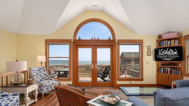 A spacious living room with a large bay window overlooking the ocean, wooden furniture, and a bookshelf. - Sea Rest: Oceanfront, private walkway to beach, hot tub, YMCA access. (Nags Head)