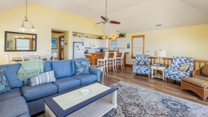 A living room with a sofa, coffee table, and patterned chairs. A kitchen with wooden cabinets and a dining area with a table and chairs is visible in the background. - Sea Rest: Oceanfront, private walkway to beach, hot tub, YMCA access. (Nags Head)
