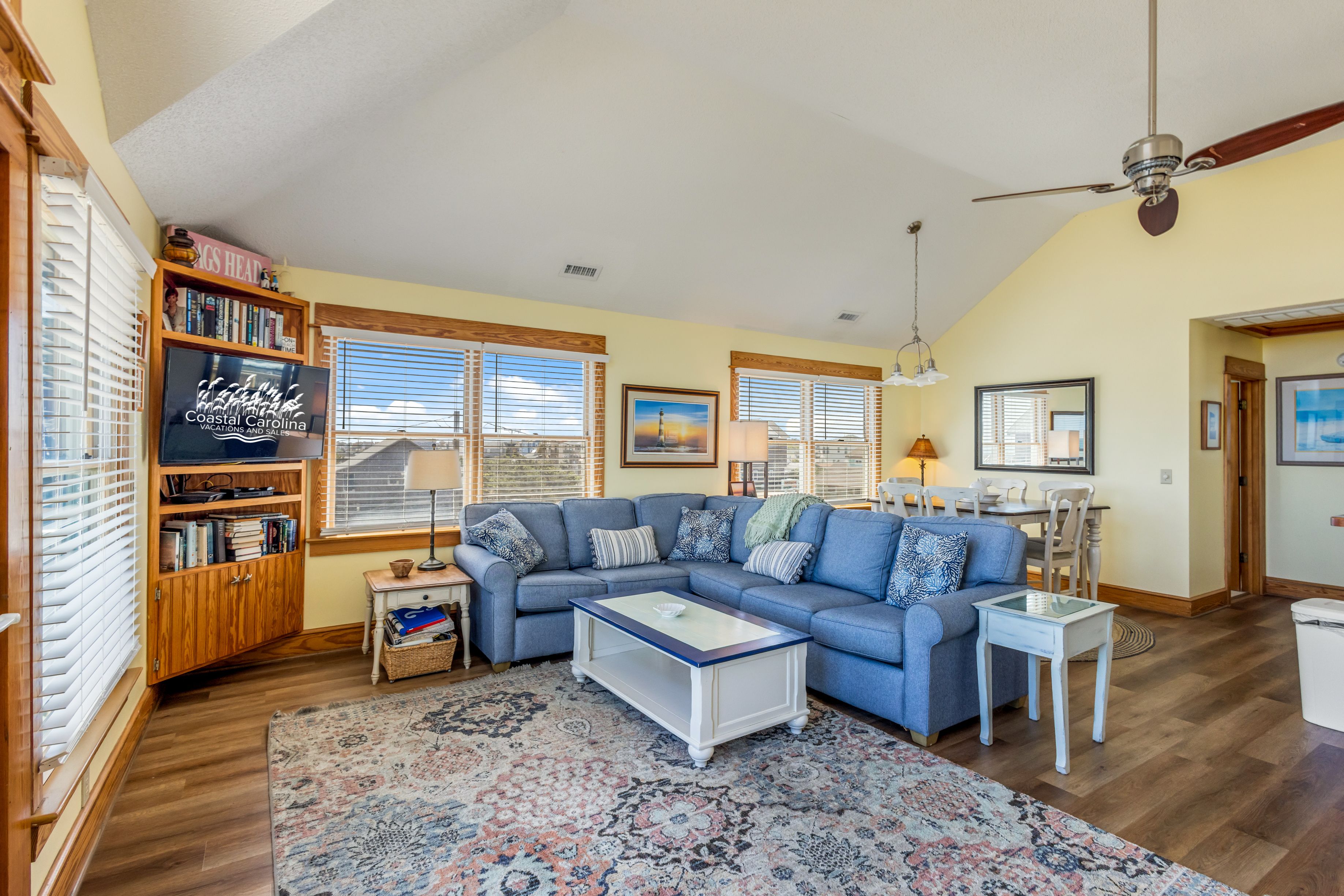 A spacious living room with a sectional sofa, a coffee table, a bookshelf, and a large window with blinds.