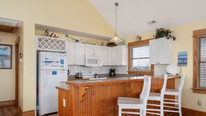 A kitchen with wooden cabinets, a white refrigerator, a microwave, and a wooden island with two white bar stools. - Sea Rest: Oceanfront, private walkway to beach, hot tub, YMCA access. (Nags Head)