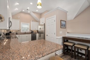 A kitchen with granite countertops, white cabinetry, a stainless steel refrigerator, and a wooden dining table with bar stools. - Calm On Down: Private pool, hot tub, elevator, dog friendly, Close to the beach! (Nags Head)