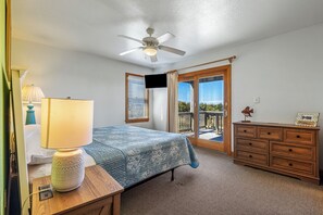 A bedroom with a bed, a desk with a lamp, a dresser, a ceiling fan, and a sliding glass door leading to a balcony.