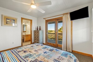 Un dormitorio con una cama grande, un ventilador de techo, una puerta corrediza de vidrio a un balcón y un televisor montado en la pared.