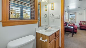 Une salle de bain équipée de toilettes, d’un lavabo et d’un meuble-lavabo. On y trouve une fenêtre avec des stores, un miroir et une horloge au mur.