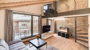 Una sala moderna con sofá, mesa de centro, televisor y chimenea. Hay un balcón con vistas al exterior.