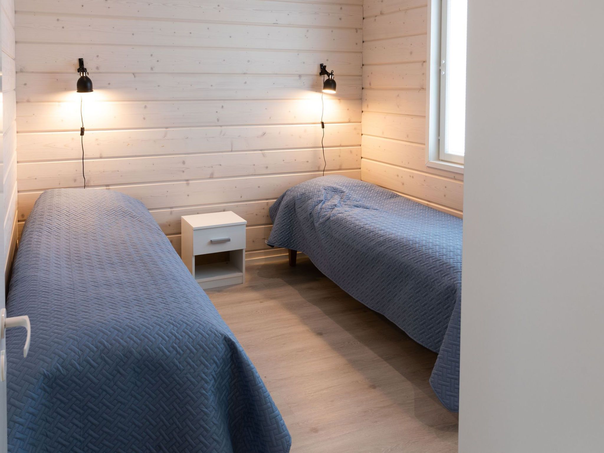 Una habitación con dos camas, una mesita de noche pequeña y blanca, y dos lámparas fijadas a la pared.