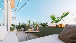 Una terrazza sul tetto con divano, un piccolo tavolo rotondo, una bottiglia e bicchieri, luci a stringa e piante in vaso.