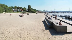 Ein sandiger Strand mit einer Holzstege, der sich in ruhiges Wasser erstreckt, umgeben von Grünflächen und Häusern.