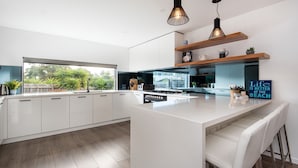 Une cuisine moderne avec des meubles blancs, un grand îlot central, des luminaires suspendus et une vue sur la verdure à l’extérieur.