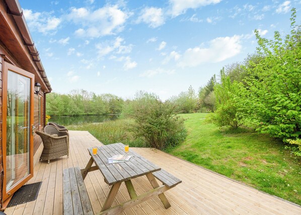 A wooden deck with a picnic table and chairs, a glass door entrance, and a view of a lake and greenery. - Stunning lodge built over the edge of the lake. (Anglesey)