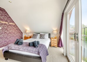 A bedroom with a bed, two bedside tables, a window with curtains, and a view of greenery outside. - Stunning lodge built over the edge of the lake. (Anglesey)