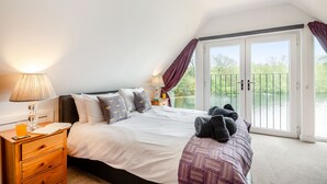 Een slaapkamer met een bed, een nachtkastje, een lamp, een raam met gordijnen en uitzicht op een waterpartij.