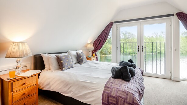 Un dormitorio con cama, mesita de noche, lámpara, ventana con cortinas y vistas a un cuerpo de agua.