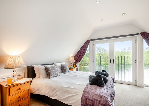 A bedroom with a bed, a nightstand, a lamp, a window with curtains, and a view of a water body. - Stunning lodge built over the edge of the lake. (Anglesey)