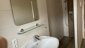 Un bagno con un lavandino bianco, uno scaffale in vetro, uno specchio e una zona doccia.